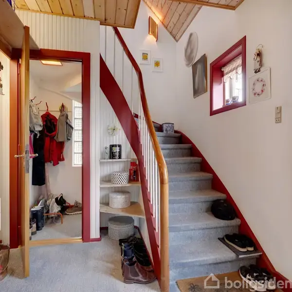Trappe snor sig opad med skridsikkert tæppe. Væggene har hvidt panel og træloft. Døre og vinduesrammer er røde. Entréen rummer sko, frakker og en åben dør til garderobe.