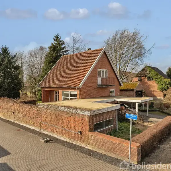 Et rødt murstenshus med skråt tag, beliggende på en stille vej omgivet af træer og buske. En carport er i forhaven, og en vejskilt viser "Røddikvej 12".