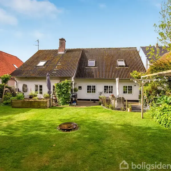 Et hvidt hus med to skorstene står i en velholdt have. Røde og brunlige tage omgiver det, mens sollys oplyser den grønne græsplæne med en lille bålplads i midten.