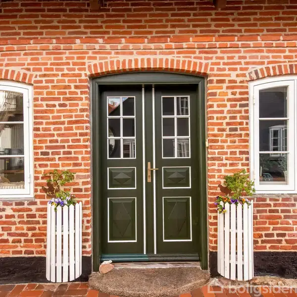 Grøn dobbeltdør med glasruder placeret mellem to hvide vinduer, omgivet af en rød murstensfacade. Foran døren står to hvide, runde plantekasser med grønne planter.