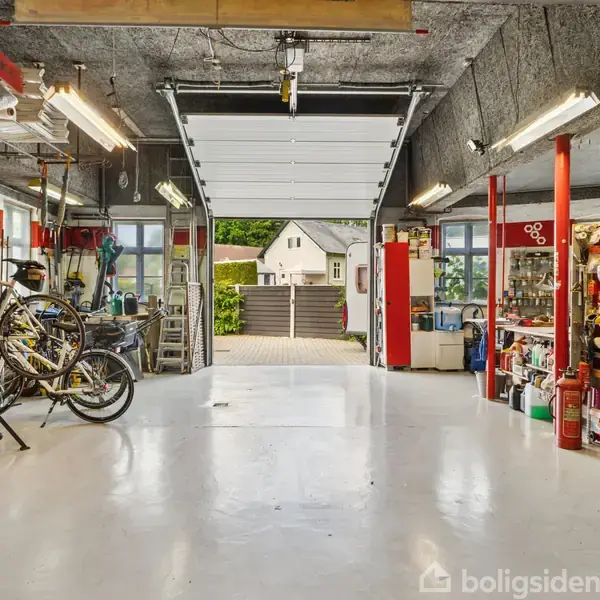 En ryddelig garage med åbneporte, cykler og værktøjsreoler. Det lyse betongulv reflekterer lysstofrør, og der er udsigt til en have og et hus i baggrunden.