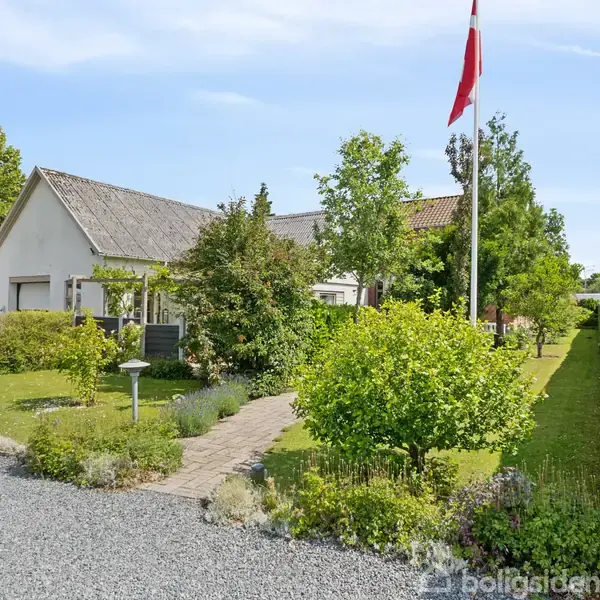 Et hvidt hus står stille omgivet af frodig have og stier. En flagstang med dansk flag vajer ved indkørslen, og en campingvogn er delvist synlig i grusopkørslen.