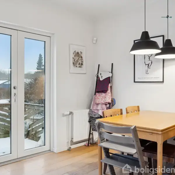 Et spisebord med fire stole står i et lyst rum. To pendellamper hænger over bordet. Glasdøre leder ud til en have med sne.