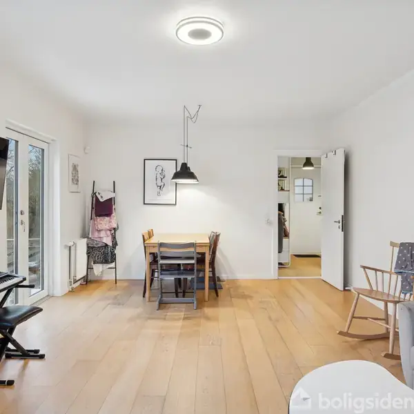 Stue med træbord og stole i midten, TV på væggen, elektrisk klaver til venstre, grå sofa til højre. Dør åbner til andet rum, lyse trægulve.