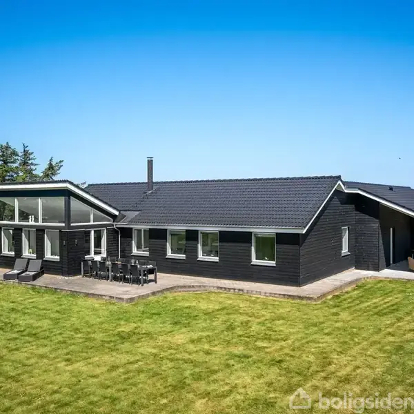 Et sort træhus står på en stor, velholdt græsplæne under en klar blå himmel. Der er et terrasseområde med havemøbler foran huset.