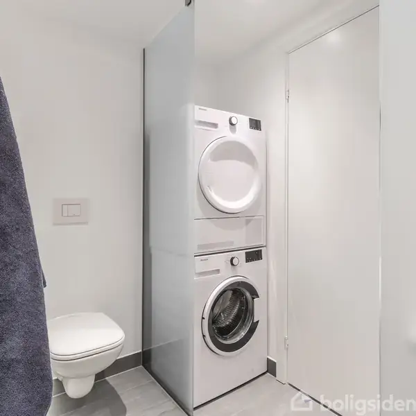 Vaskemaskine og tørretumbler er stablet i et moderne badeværelse; ved siden af hænger et mørkeblåt håndklæde ved et vægmonteret toilet. En enkel, hvid dør ses i baggrunden.