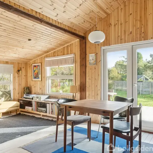 Et spisebord med to stole står i et rum med trævægge og loft. Store vinduer og glasdør lukker lys ind og giver udsigt til en have.
