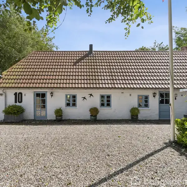 Et hvidt, langstrakt hus med teglstenstag og seks vinduer ligger foran en grusplads. Plantekrukker pynter facaden, og tallet "10" er på muren. Omkranset af grøn natur.