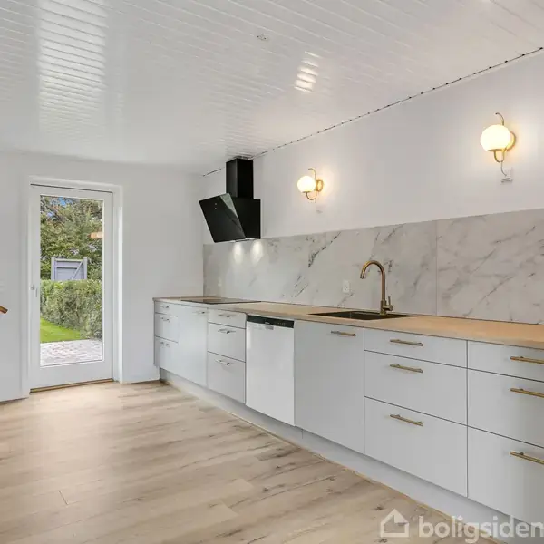 Et moderne køkken med hvide skabe og marmor-bagside, indeholder en sort emhætte og indbygget ovn. En åben dør fører ud til en have. Til venstre ses en trappe.