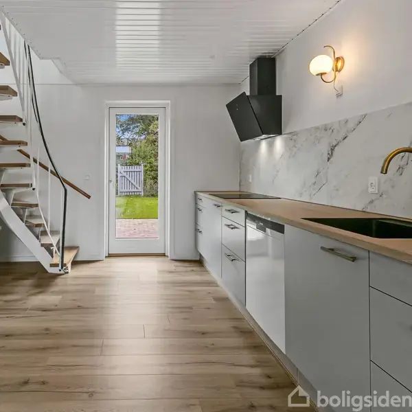 Køkken med hvide skabe og marmorvæg, indeholder vask og emhætte. Trappe til venstre fører opad. En åben dør viser have med græsplæne og stakit i baggrunden.