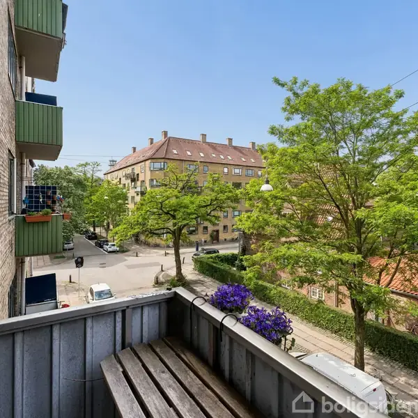 En balkon med en træbænk er placeret i en urban gade udstyret med træer. Et skilt med teksten "REAL TIL SALG" og et telefonnummer hænger på bygningen.