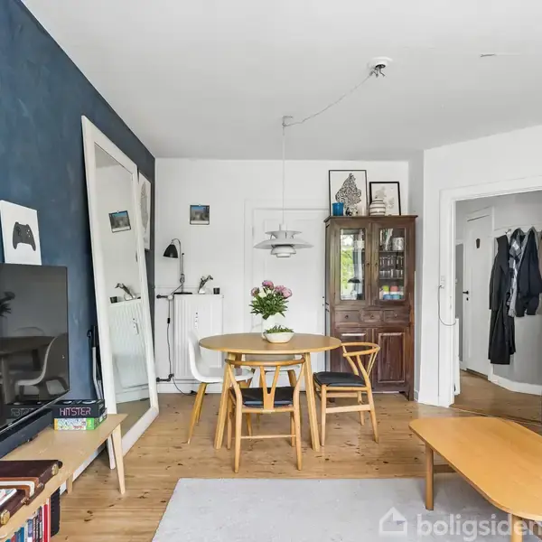 En stue med et rundt spisebord og stole i træ i midten. En sofa står til højre, og et fjernsyn er monteret på en mørkeblå væg til venstre.