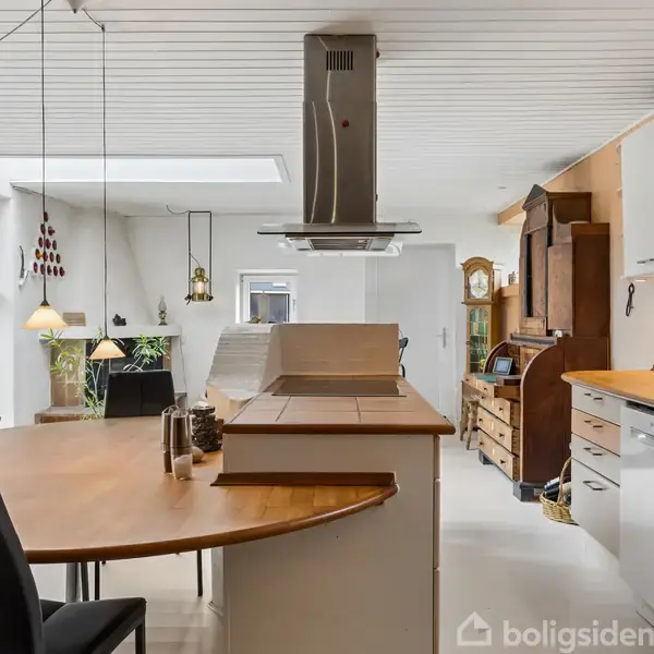 Køkken med træbordplader, emhætte over kogeø, og siddepladser. Baggrunden viser en stue med sofa, planter, og dekorative genstande. Lyse vægge og loft giver et rummeligt indtryk.