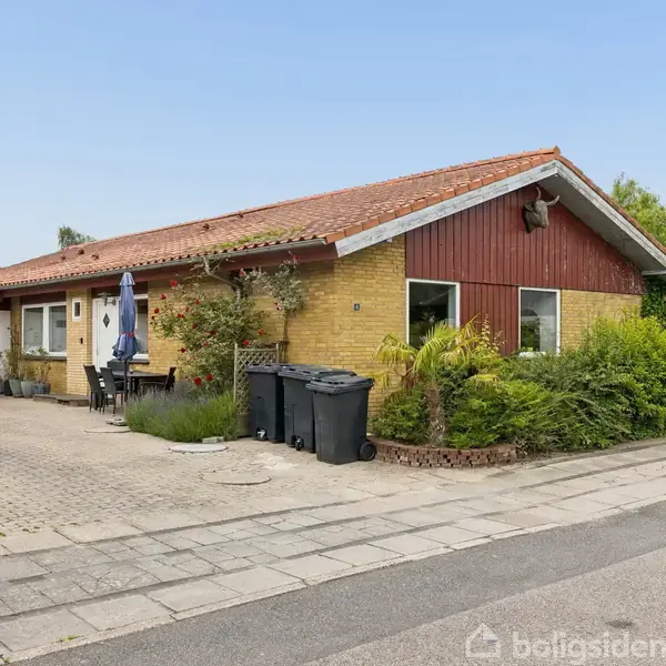 Et gult murstenshus med rødt tag, garage til venstre og terrasse foran. Fire skraldespande står foran huset, omgivet af buske og planter langs gangstien.