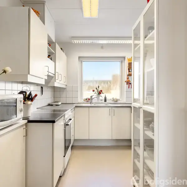 Et moderne køkken med hvide skabe og apparater. Der er en mikrobølgeovn på bordet. Et stort vindue bagerst giver naturligt lys. Hylder indeholder diverse genstande.