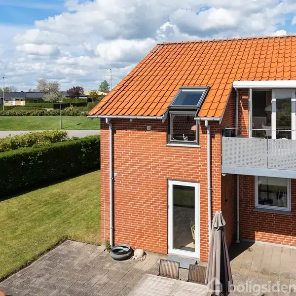 Hus med rødt tegltag og en altan. Stående i en solrig, grøn have med en flisebelagt terrasse omkranset af hække. Skyet blå himmel i baggrunden.