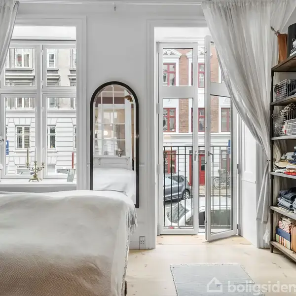 Et soveværelse med en seng står ved siden af et åbent fransk vindue, som fører ud til en lille altan. Gardinerne er trukket tilbage, og en åben reol viser tøj.