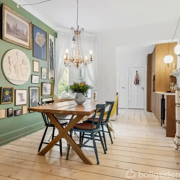 Et spisebord med stole står på et trægulv i et rum med grøn væg dækket af kunstværker. En lysekrone hænger over bordet. Et skab og komfur ses i baggrunden.