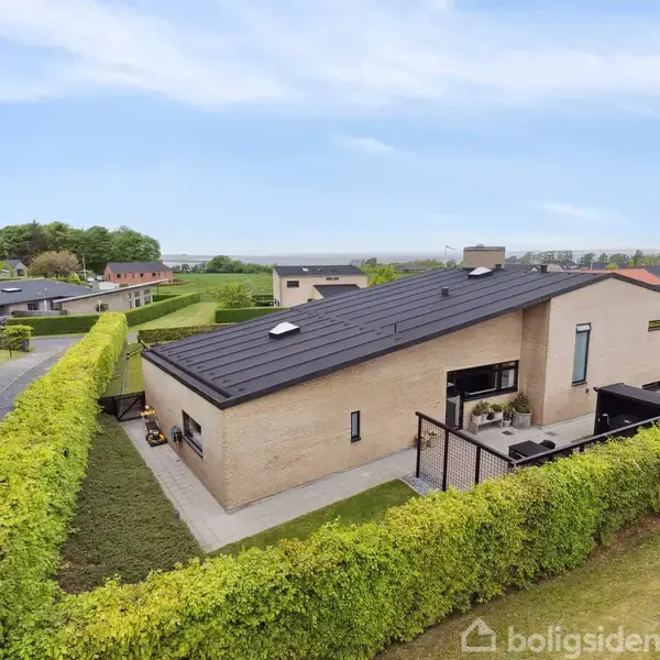 Moderne, ensidig taghus med store vinduer ligger omgivet af tætte hække i et boligkvarter. Den omgivende have er velplejet med en terrasse og græsplæne.