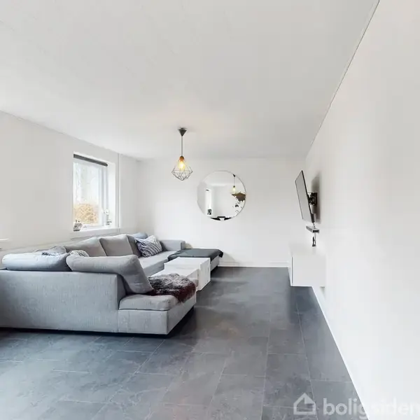 Grå sofa står mod en hvid væg i stue. Fladskærms-TV er monteret på modsatte væg. Rummet har store vinduer og mørkegråt gulv. Rundt spejl hænger i baggrunden.