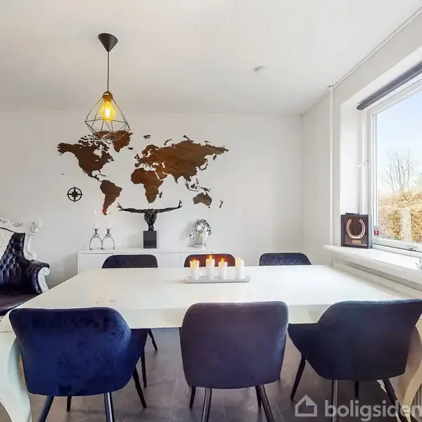 Et hvidt spisebord er omgivet af blå stole. Over bordet hænger en lysekrone. På væggen bagved er et verdenskort i brun. Et vindue viser grønne buske udenfor.