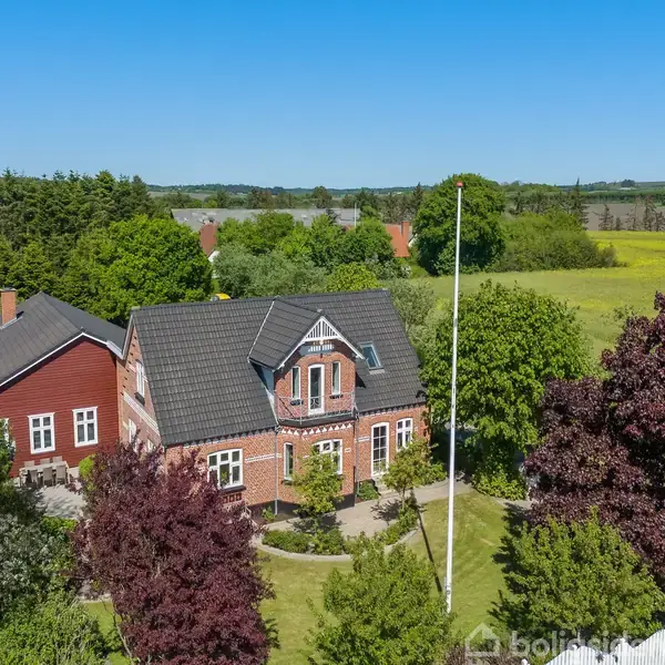 Et rødt murstenshus med gråt tag står omgivet af frodig have med træer i forskellige farver. Baggrunden består af åbne marker og skov i klart solskin.