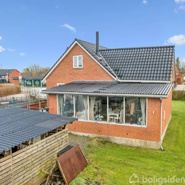 Rødt murstenshus med sort tag, stor glasoverdækket veranda fyldt med møbler. Huset ligger i en villavej med græsplæne omkring det. Flere huse kan ses i baggrunden.