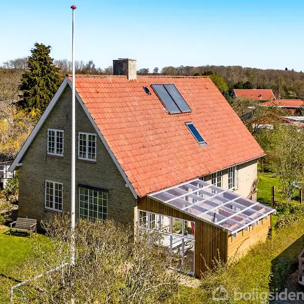Et hus med rødt tegltag har en glasudestue i haven omkranset af græs og træer i en forstad. Flagstangen står ved siden af huset.
