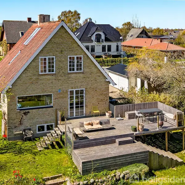 Et murstenshus med rødt tag har en stor træterrasse med siddepladser. Huset ligger i en have med græs og træer i et beboelsesområde med flere lignende huse.