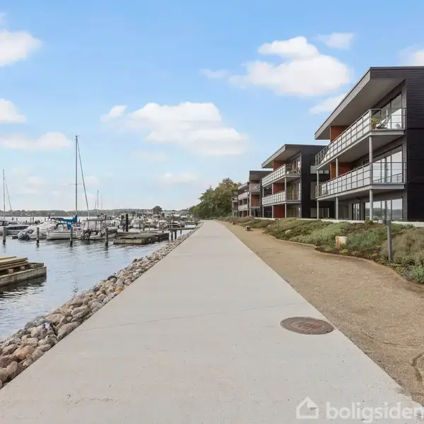 Bygningskompleks løber langs en havnepromenade med fortov, hvor lystbåde ligger fortøjet i vandet. Skyfri himmel og grøn bevoksning ved siden af bygningerne.