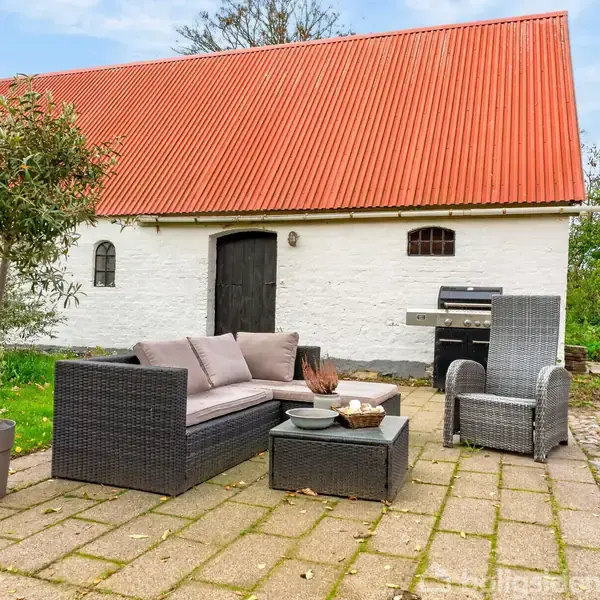 Et udendørs siddeområde med polyrattanmøbler står på en terrasse. En potteplante og et grillområde er nærved. Baggrunden viser en traditionel lade med hvid væg og rød tag.