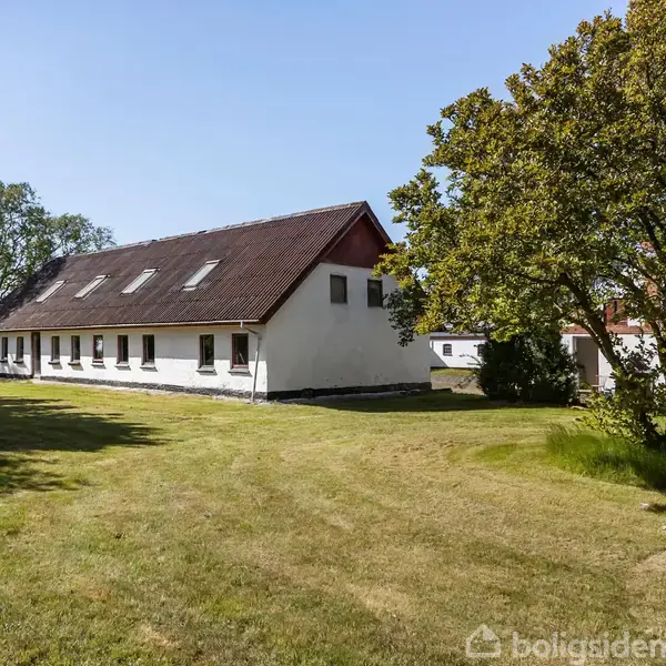 Et langt, hvidt hus med en mørk tagetage står stille på en velholdt, grøn græsplæne. Store træer omgiver bygningen, der har flere vinduer og en rolig atmosfære.