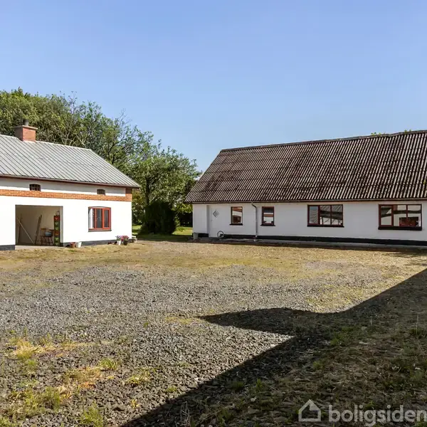 To bygninger står omkring en åben gårdsplads med grus. Den ene bygning har en garage med en bil og åbne døre. Træer omkranser området under en blå himmel.