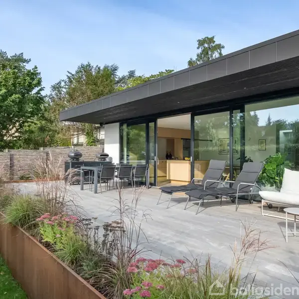 Moderne hus med glasfacade åbner ud til en terrasse med havemøbler. Omgivet af velplejet have med græsplæne og blomsterbede, skaber det en fredfyldt, naturlig atmosfære.