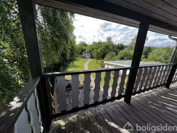 Balkon med trægulve, udsigt til en have med en lille sti, der fører til et hvidt hus. Omgivet af frodig vegetation og træer.