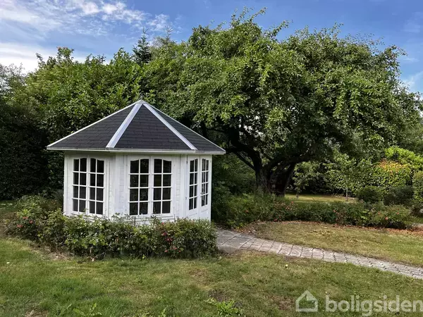 Et hvidt havepavillon står stille i en grøn have, omgivet af blomster og græs. En stor trækrone kaster skygger i baggrunden under en blå himmel.