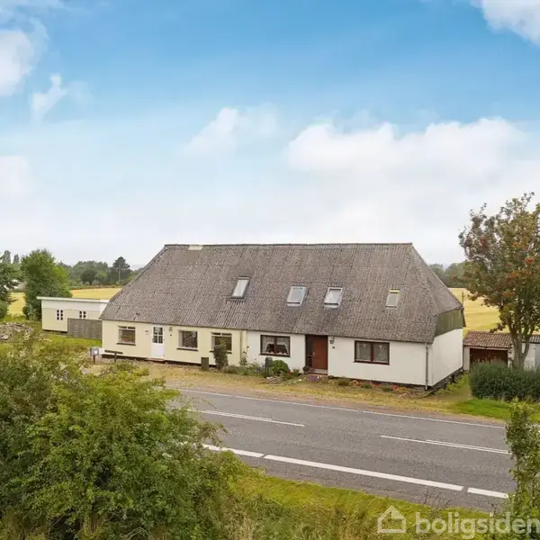 Et hvidt hus med stråtag ligger ved en vej omgivet af landlige marker og træer. Huset har flere vinduer og en rød dør, og himlen er let overskyet.