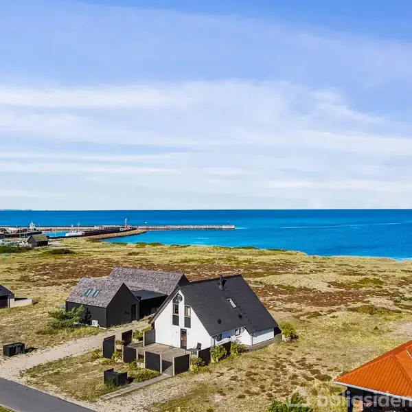 Hus med skifergråt tag står på en græsklædt grund ved kysten. Det ligger tæt på andre huse og en lystbådehavn, mens det klare, blå hav strækker sig i baggrunden.