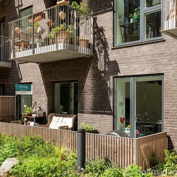 En stuelejlighed med åbent skydedørsvindue og en lille terrasse omgivet af en lav træhegn. Ovenfor er en balkon med planter og potter. Et skilt viser "Til salg". Grønt område omkranser bygningen.