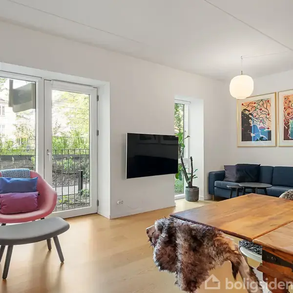 Stuen indeholder en pink stol med farvede puder, placeret ved siden af en åben dør mod haven. En sofa står i hjørnet, dekoreret med kunst på væggen. En hængende lampe og et fjernsyn på væggen ses også.