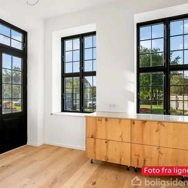 Et lyst rum med store vinduer og en dør lavet af sort træ og glas. En trækommode står på et lyst trægulv. Tekst: “Foto fra lignende bolig”.