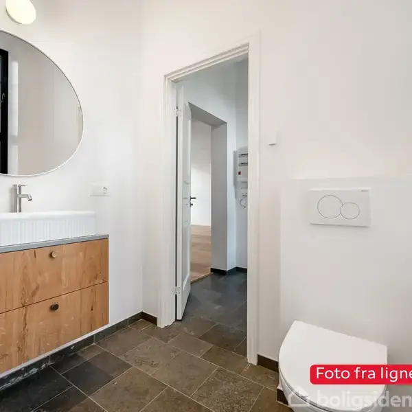 Ovalt spejl hænger over en hvid vask på et lyst træskab i et badeværelse med sorte fliser. En dør fører til en anden rum. Tekst: "Foto fra lignende bolig".