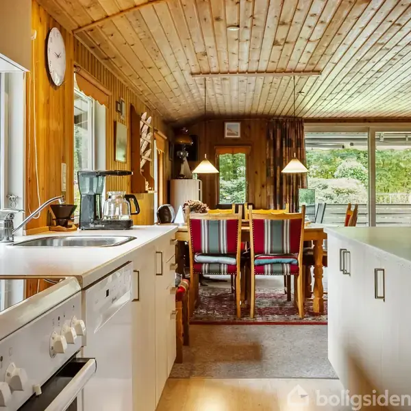 Køkken med hvidt kabinet og vask, kaffemaskine ved vinduet. Spisebord og stole i træ ligger ved siden af store vinduer, der viser en frodig, grøn have.