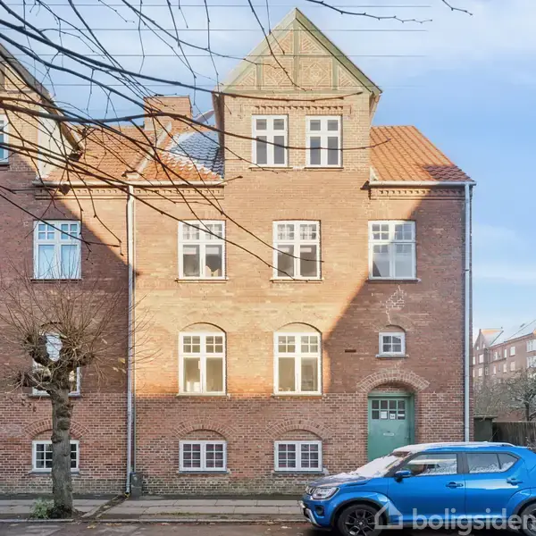 Bygning i mursten med spidse tage står på en stille gade. En blå bil er parkeret foran, og et trafikskilt ses til højre. Himlen er klar og blå.