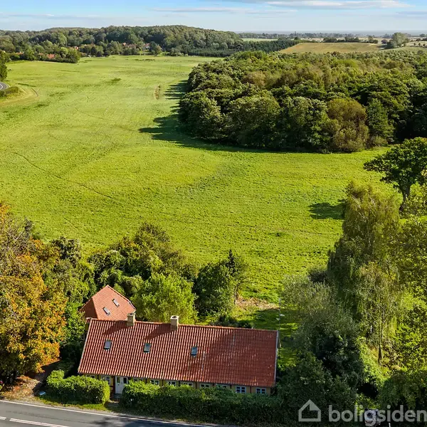 Hus med rødt tegltag ligger omgivet af træer ved en landevej. Baggrunden består af en stor grøn mark og skovklædte bakker i horisonten.