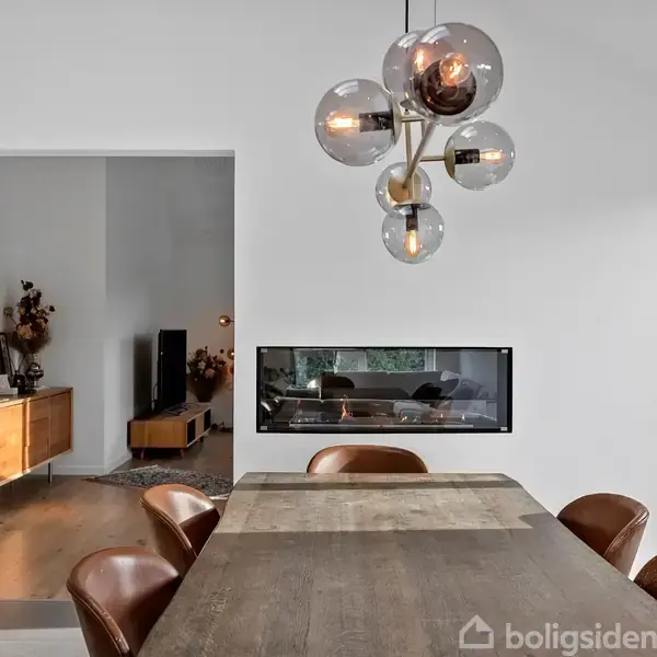 En spisestue med et træbord omgivet af læderstole; en moderne lysekrone hænger over bordet. Bagved ses en gennemgående pejs og en stue med en kommode og dekorationer.