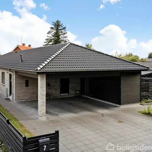 Et moderne, lavt etplanshus med fladt tegltag står stille. Der er en indkørsel foran med fliser, en carport til venstre og en sort låge med tallet "7".