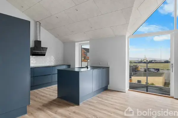 Et moderne køkken med mørkeblå skabe og lys træparketgulv, står i et rum med store vinduer, der giver udsigt til en åben himmel og et kystområde udenfor.