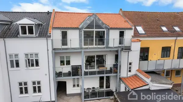 En treetagers bygning med flere altaner har en rød tegltag og moderne glasdetaljer. Bygningen er omgivet af nabohuse under en blå, let skyet himmel.