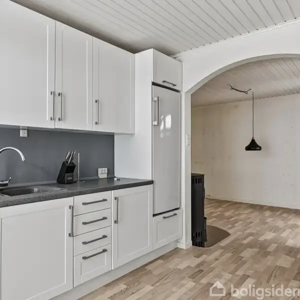 Hvidt køkken med hvide skabe og en grå bordplade, hvor en kaffemaskine er placeret. Køkkenet fører gennem en bue til et rum med lys træparket og en hængende sort lampe.